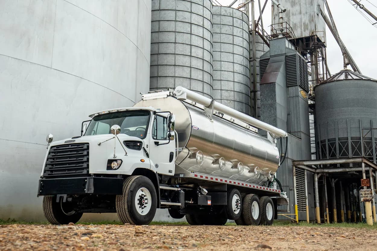 Ledwell stainless bulk feed body parked beside grain silos